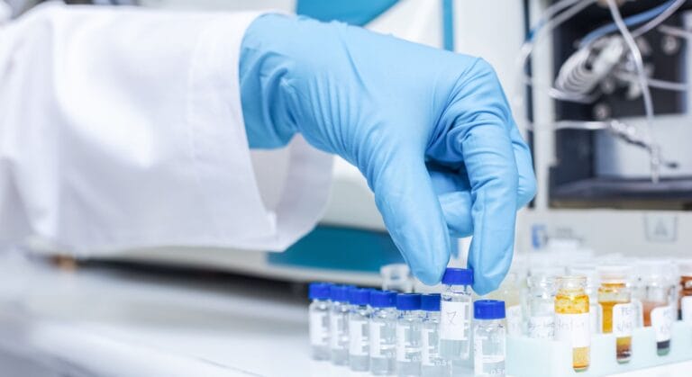up to data StudyGen 360 StudyLab | Close-up of a gloved hand in blue latex gloves holding a small glass vial with a blue cap in a laboratory setting, with additional sample vials containing various colored liquids visible in the background alongside white laboratory equipment and instruments.